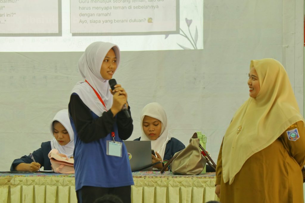 Young woman in hijab speaks into microphone, another woman listens.